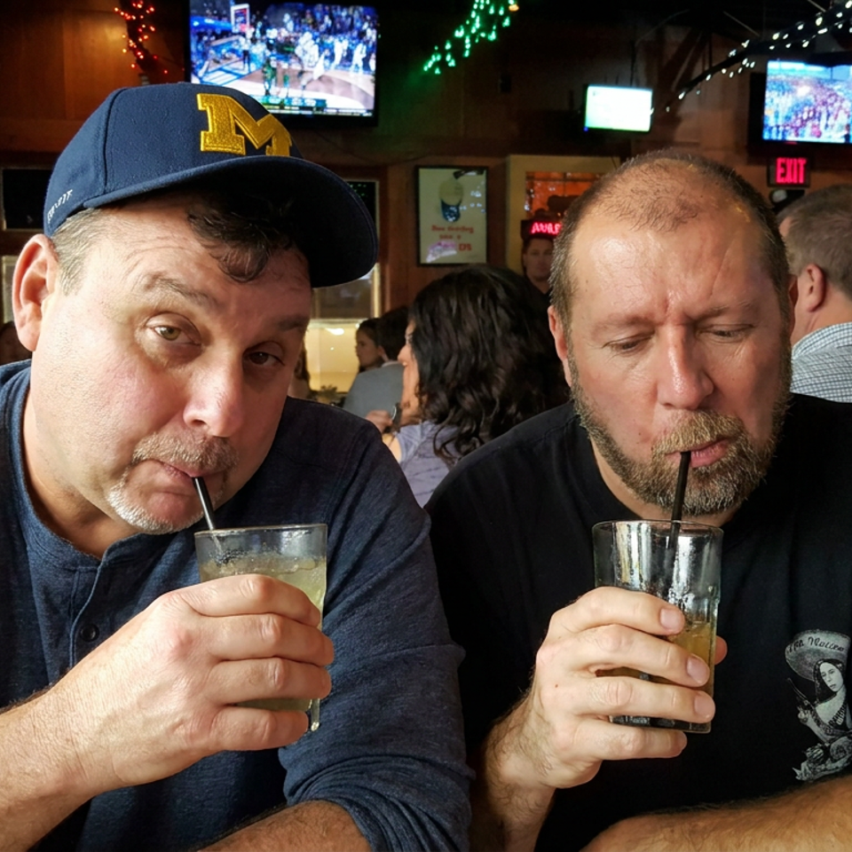 Two men sip drinks at a lively sports bar surrounded by televisions, patrons, and colorful string lights.