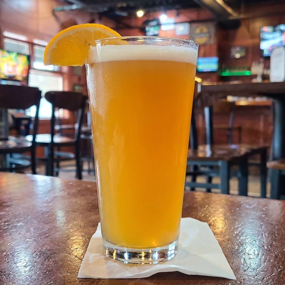 Pint of golden beer garnished with an orange slice on a napkin at a warmly lit sports bar.