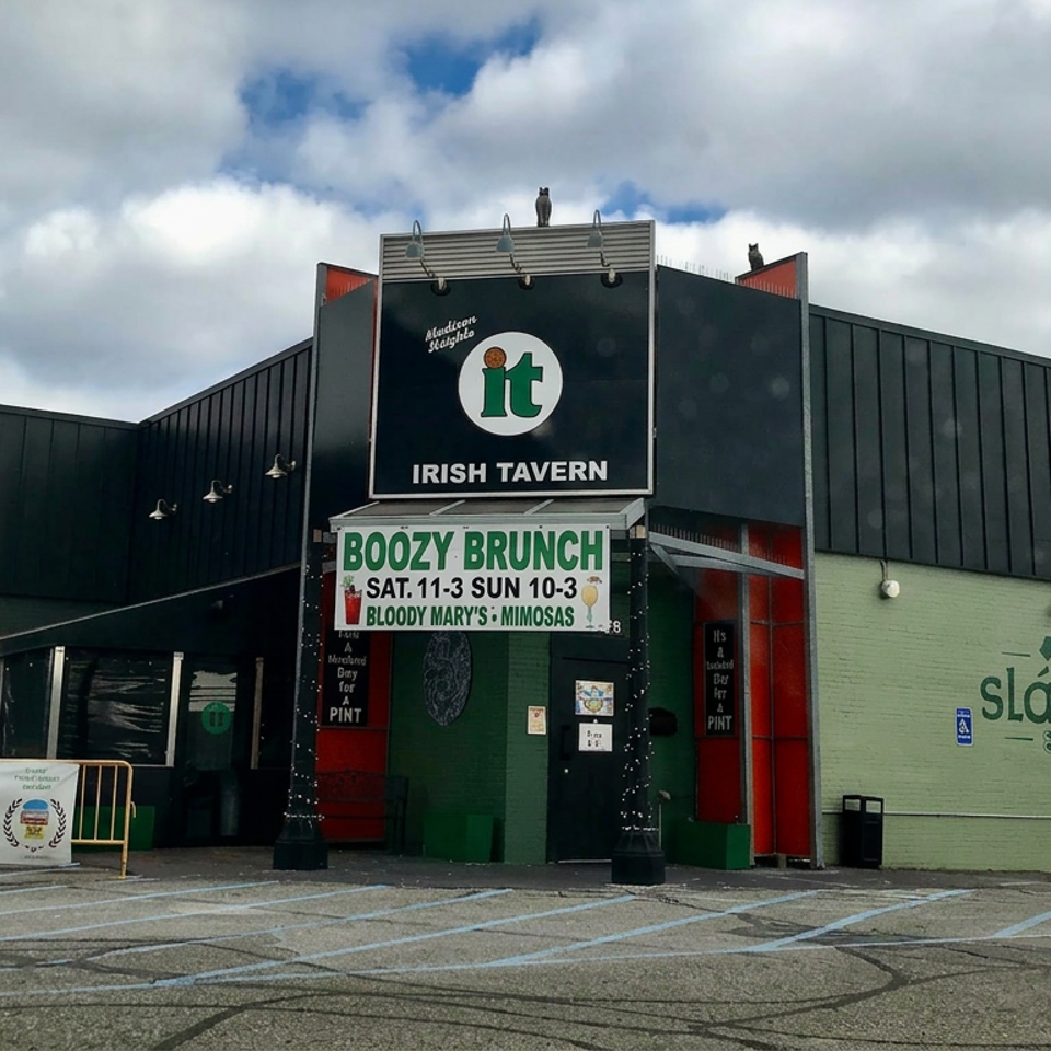 Exterior of an Irish tavern with green branding, brunch signage, mural, and a mostly empty parking lot under cloudy skies.