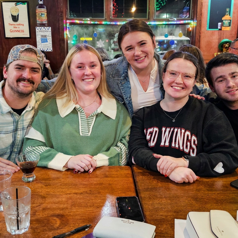 Five friends smile together at a wooden restaurant table surrounded by drinks, arcade machines, and festive decorations.