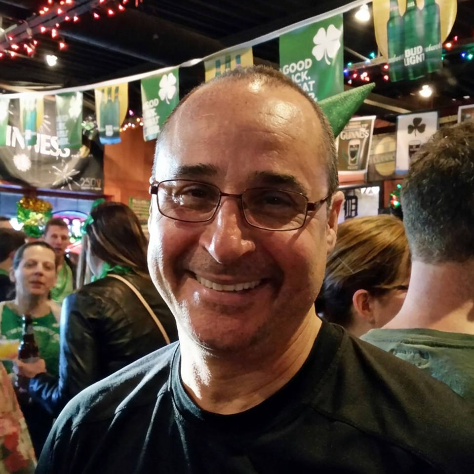 Smiling man wearing glasses at a crowded bar decorated with green St. Patrick’s Day banners and string lights.